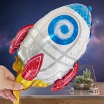 O mana tinand un balon din folie metalizata in forma de racheta spatiala alba cu detalii rosii albastre si flacari aurii deasupra unei mese