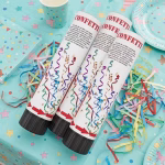 Trei tuburi cu confetti asezate pe o fata de masa albastra decorata cu stelute alaturi de fasii colorate si pahare de petrecere