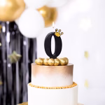 Lumanare festiva pentru tort in forma cifrei zero de culoare neagra avand aplicata o coronita aurie cu sclipici in partea dreapta sus.