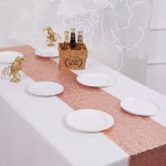 Decor de nunta la malul marii cu o masa alba decorata cu o traversa roz auriu stralucitoare, un tort supraetajat si flori.