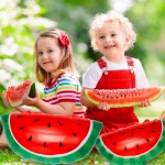 Doi copii zambitori stand pe iarba verde mancand pepene natural langa doua baloane din folie in forma de felie de pepene