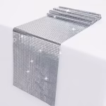 Traversa De Masa Cu Paiete Argintii Traversa de masa argintie decorata cu paiete patrate stralucitoare asezata pe o fata de masa alba.