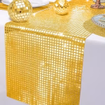 Traversa De Masa Cu Paiete Aurii Traversa de masa aurie decorata cu paiete patrate stralucitoare asezata pe o fata de masa alba langa doua globuri disco aurii.