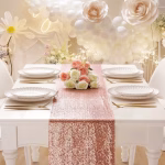 Traversa De Masa Cu Paiete Roz Auriu Rose Gold Masa festiva decorata cu o traversa cu paiete roz auriu, farfurii albe, tacamuri aurii, flori si o arcada din baloane albe in fundal.