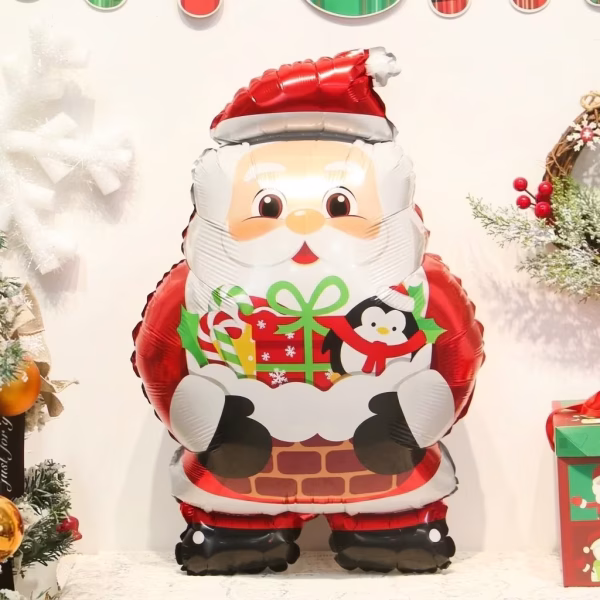 Balon din folie de 66 cm in forma de Mos Craciun iesind dintr-un semineu de caramida, tinand in brate un cadou impachetat festiv si un pinguin simpatic