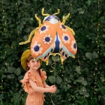 Balon din folie in forma de buburuza avand aripile de culoare corai decorate cu flori roz si albastre contururi aurii stralucitoare antene aurii si un corp de culoare bleumarin