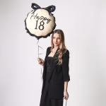 Balon din folie in forma de medalion pentru majorat avand un fundal crem inconjurat de o margine neagra ondulata decorat in partea superioara cu o funda neagra si textul caligrafic Happy 18 in centru