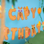 Banner de petrecere format din litere groase de culoare maro deschis care compun mesajul CAPY BIRTHDAY decorate haios cu ochelari de soare coifuri de petrecere portocale o ratusca si flancate de doua figurine capybara zburand cu baloane
