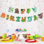 Banner din carton cu mesajul Happy Birthday, format din litere colorate decorate creativ sub formă de dinozauri simpatici, incluzând un pui care iese din ou, dinozauri cu coarne și creaturi zburătoare, în nuanțe de verde, portocaliu, maro și albastru