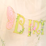 Banner de petrecere cu mesajul Happy Birthday format din litere roz si verzi decorate cu liane flori de camp ciupercute rosii si zane delicate cu aripi