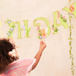 Banner de petrecere cu mesajul Happy Birthday format din litere roz si verzi decorate cu liane flori de camp ciupercute rosii si zane delicate cu aripi