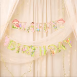 Banner de petrecere cu mesajul Happy Birthday format din litere roz si verzi decorate cu liane flori de camp ciupercute rosii si zane delicate cu aripi