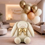 Balon din folie tridimensional in forma de iepuras crem cu obraji roz si funda aurie voluminoasa la gat asezat pe podea acompaniat de un buchet de baloane din latex umflate cu heliu in nuante de auriu chrome bej si alb nisip