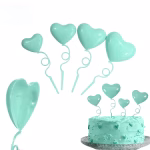 Set de 4 toppere din plastic lucios in forma de inimi tip balon de culoare turcoaz, prevazute cu tije ondulate ce imita panglicile, asezate ca decor pe un tort asortat in aceeasi nuanta
