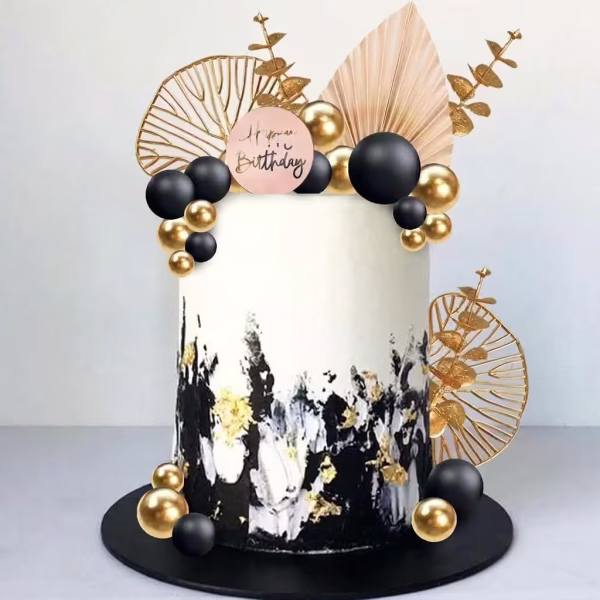 Tort alb cu texturi negre si aurii, decorat in varf si la baza cu un aranjament spectaculos de sfere negre si aurii, frunze evantai bej, crengute aurii inalte si un topper rotund Happy Birthday