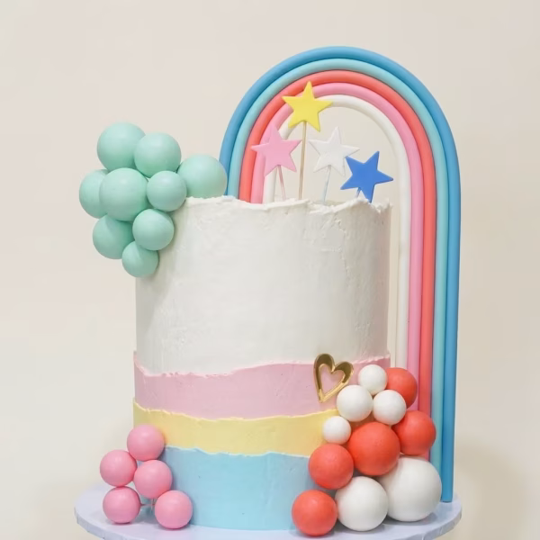 Tort alb cu straturi pastelate decorat cu un curcubeu gigantic, acompaniat de stele decupate din spuma pe bat si grupuri de sfere mate in culorile menta, roz, coral si alb